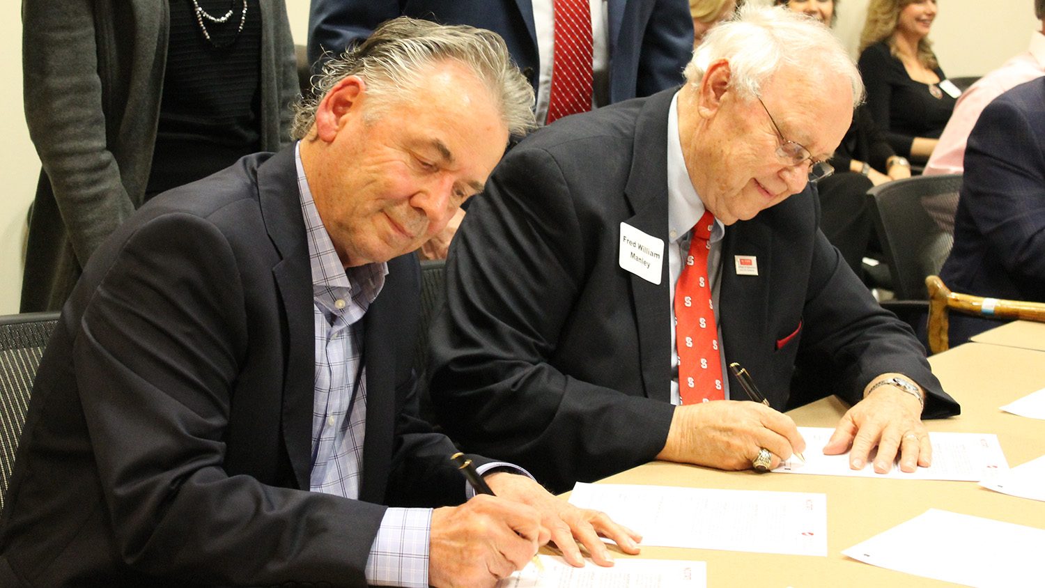 Two men seated at a table, signing papers.