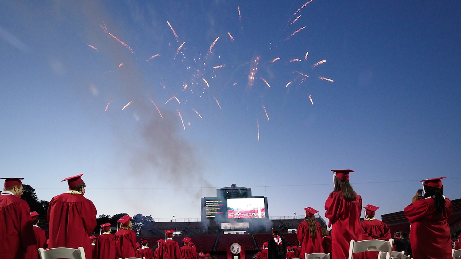 Students in graduation robes watch fireworks at Carter-Finley Stadium during the Spring 2021 Commencement
