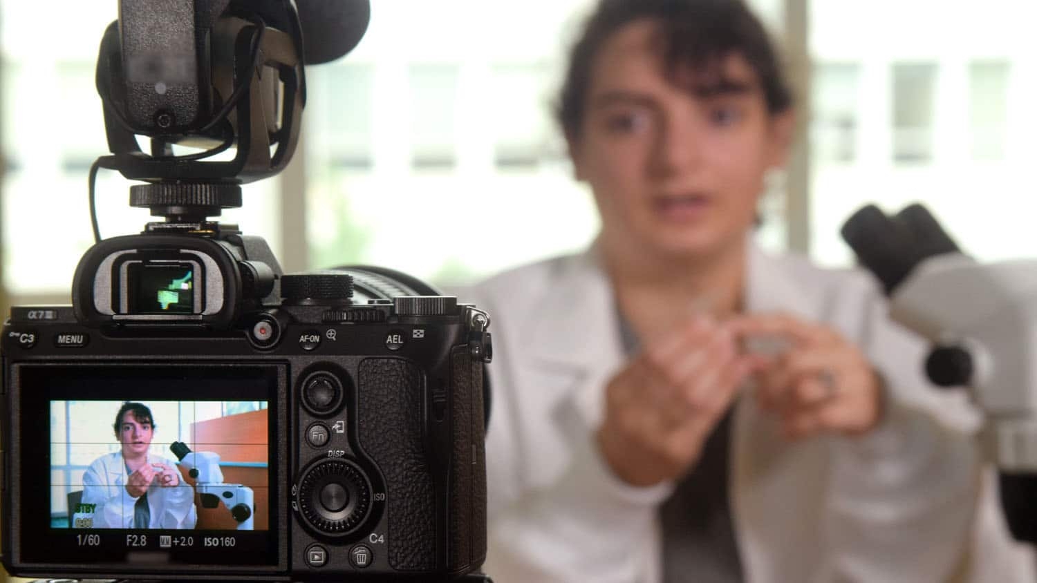 a camera is filming a scientist talking about her work; the scientist is also visible in the viewfinder of the camera