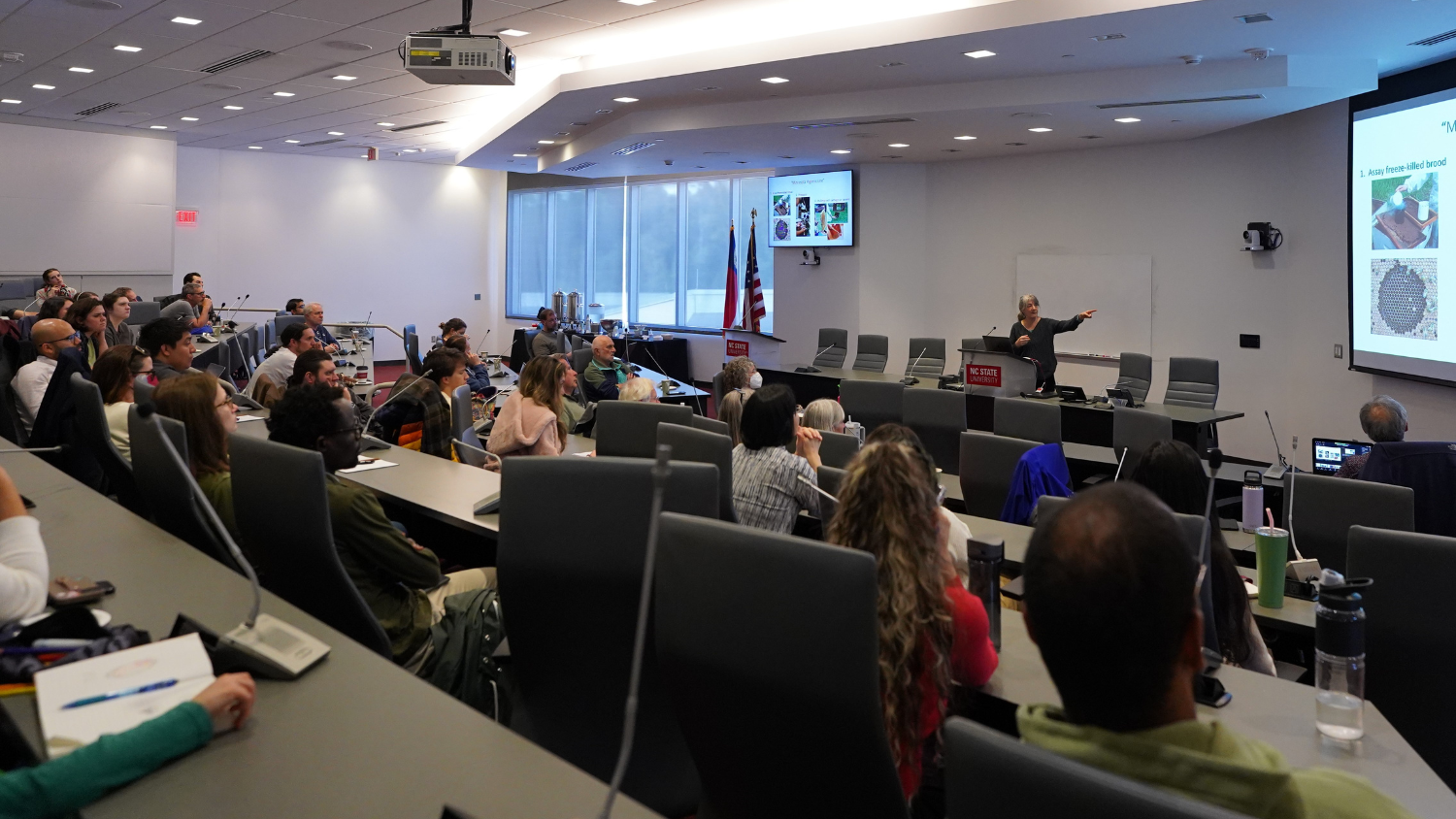 Viewed from the back of a lecture hall, an audience listens to a seminar with the speaker at the front of the room in front of a projector screen