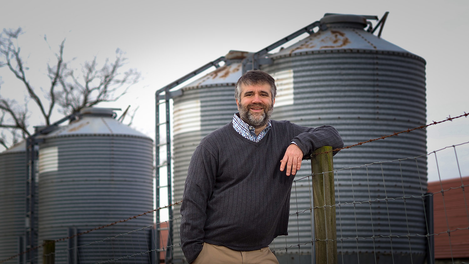 Man leaning against a fence post, with grain bins in the background.