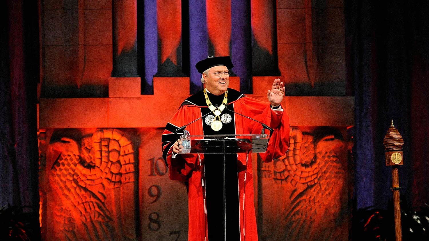 Chancellor Woodson wearing red commencement regalia, standing at a podium with an image of the Belltower in the background, and waving to the crowd