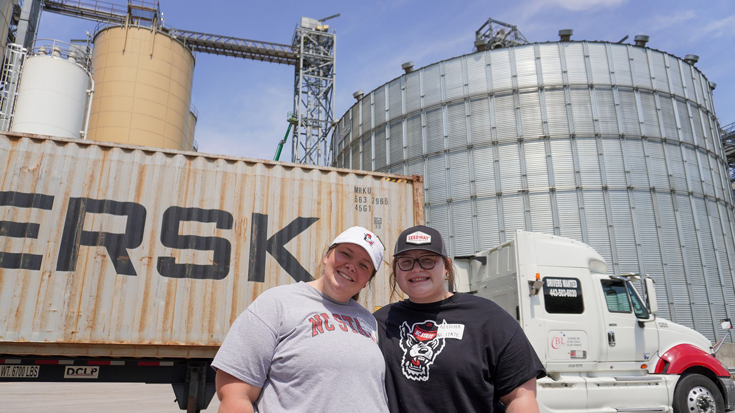 two women stand in front of a mac truck and grain silo