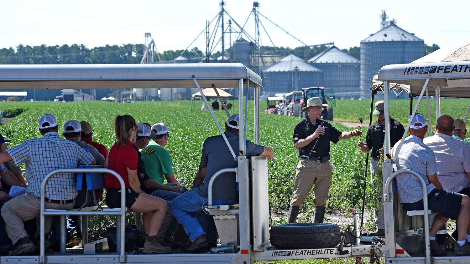 Extension specialist giving a field demonstration in a soybean field while onlookers in a tram observe.