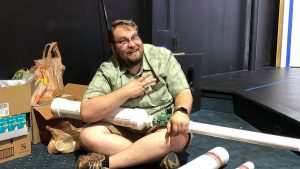 White male sitting on the floor holding PVC piping. 