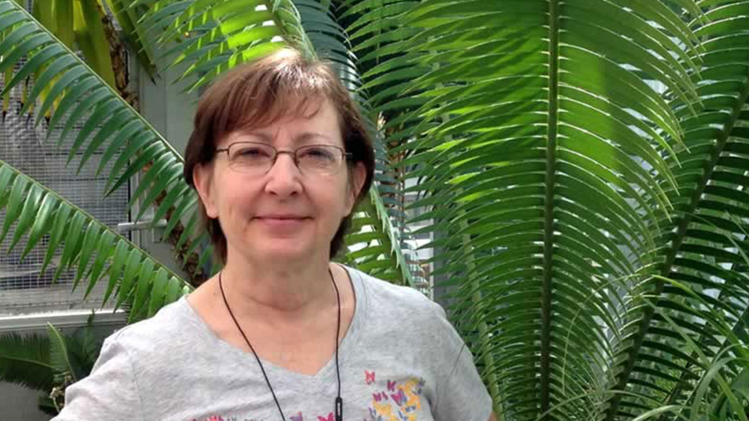 White woman with glasses with large fern in the background.