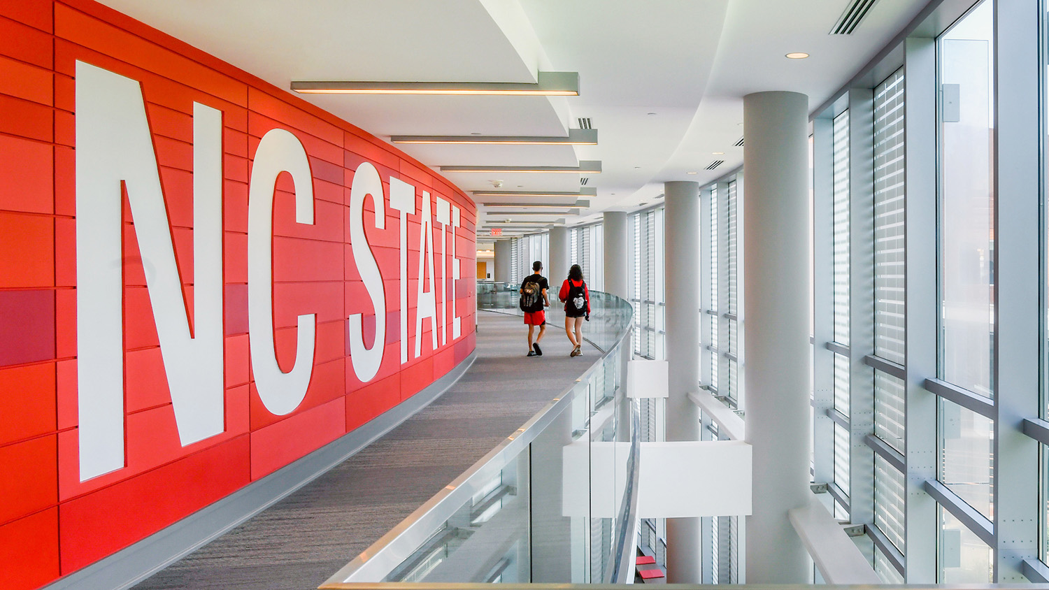 Two people walking along a glass hallway with large NC State mural.