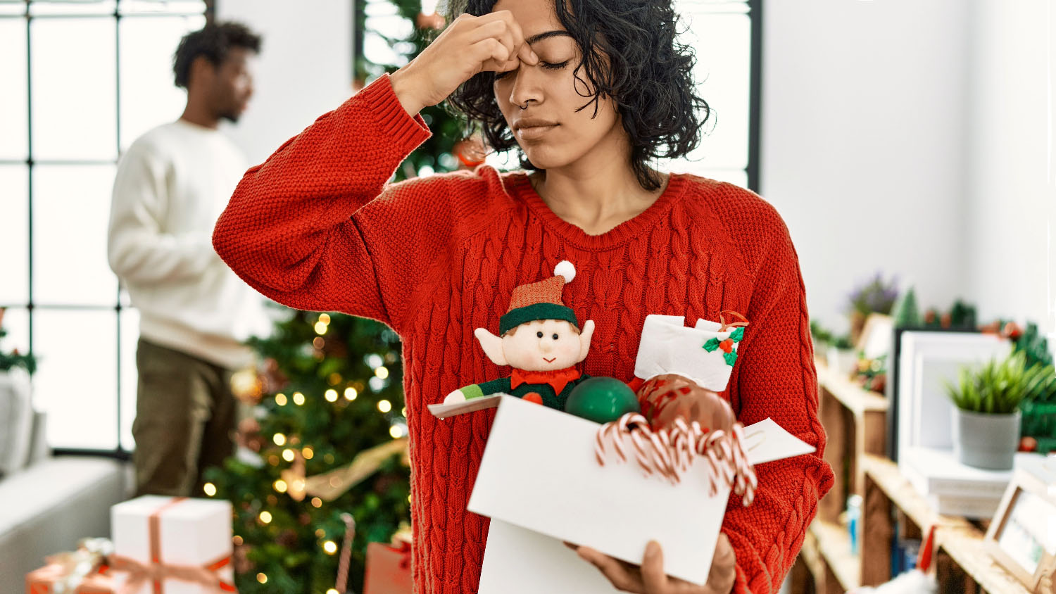 woman wearing a red sweater pinches her eyebrows while a man in the background decorates a christmas tree