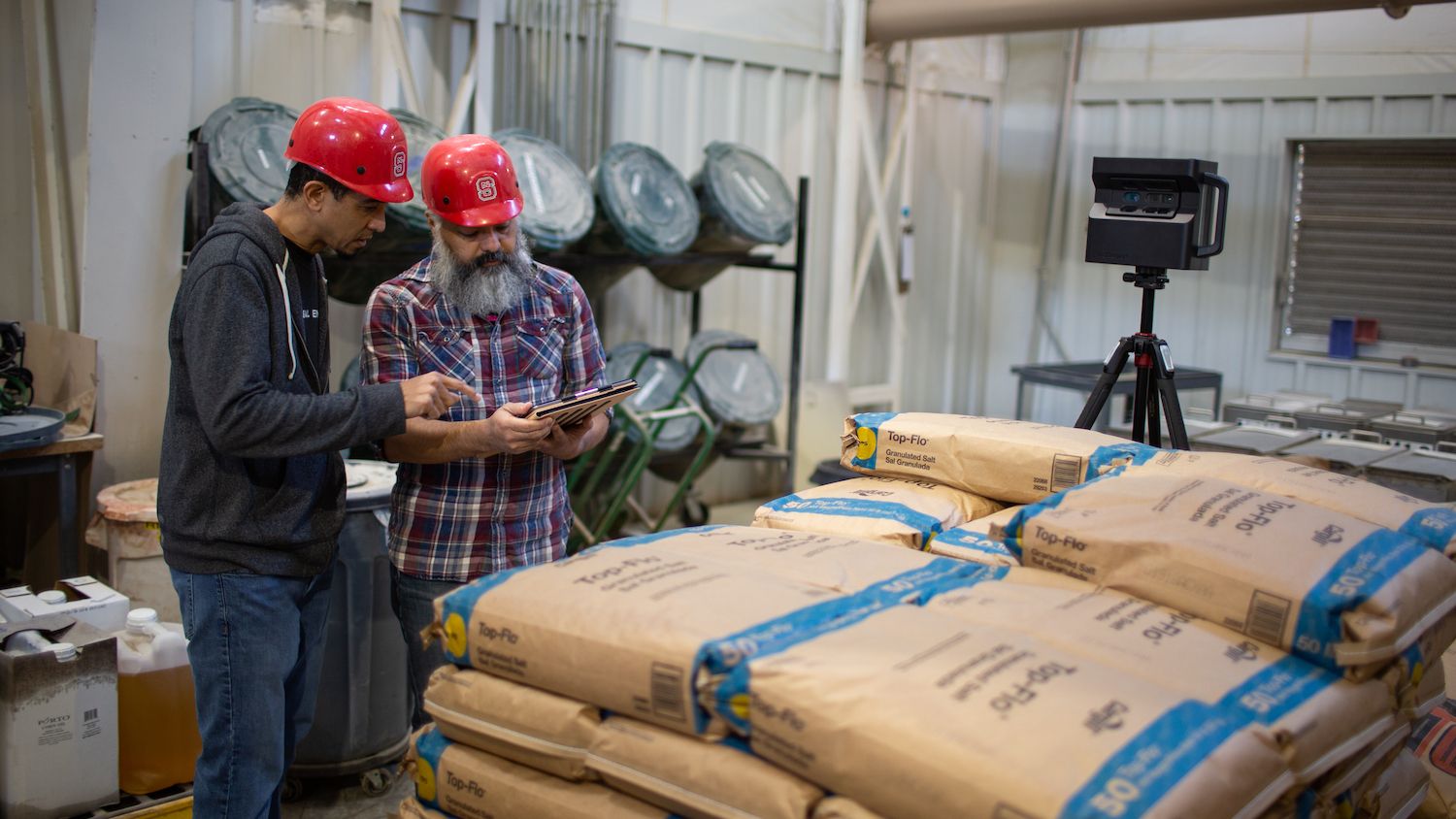 Two workers in red hard hats stand near stacked bags of animal feed