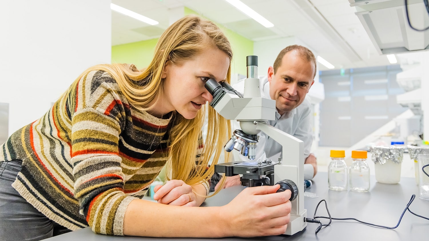 Postdoctoral researcher Lisa Van den Broeck looks into a microscope while STEPS Center director Jacob Jones watches on by her side..
