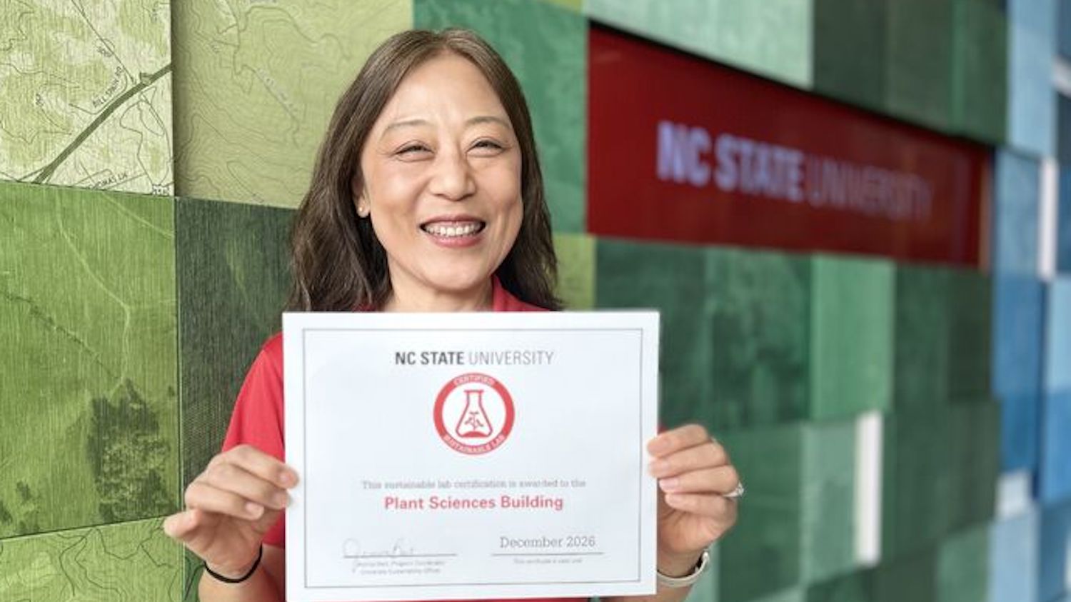 woman holds a certificate in front of a wall with the NC State University logo