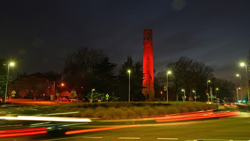 NC State University's belltower, lit in celebratory red as vehicles pass through a roundabout at night.