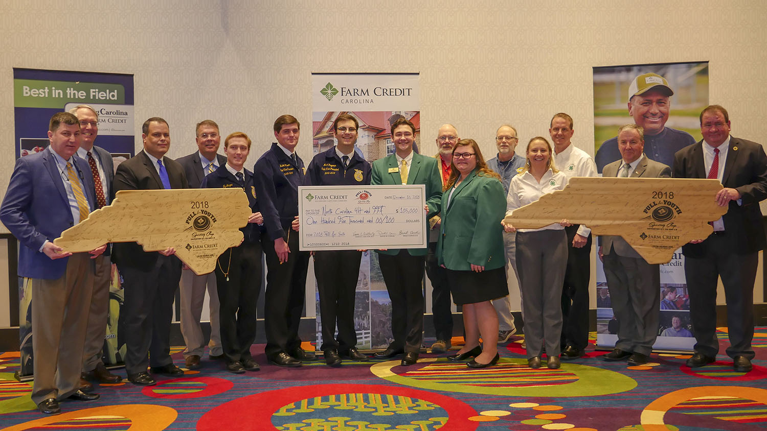 15 people holding a check for $105,000 and two large wooden plaques in the shape of North Carolina