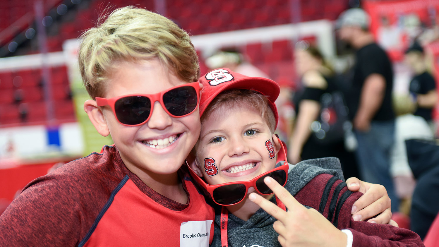 Two young boys, one in red shirt with red sun glasses, the other with a red NC State hat, red sunglasses and State tattoos on cheeks.