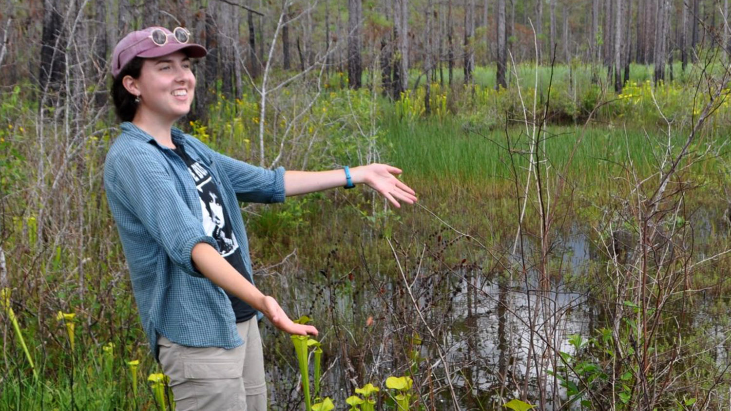 A scientist stands in a swamp, arms outstretched, looking happy.