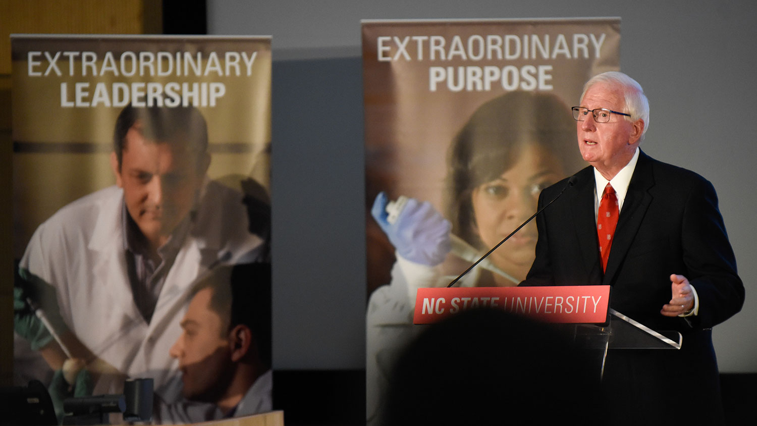 Man at podium with banners behind him that say "Extraordinary Leadership" and "Extraordinary Purpose"