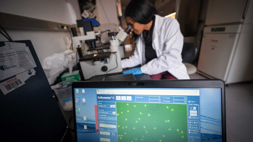 a computer screen with data and a female research looking through a microscope in the background
