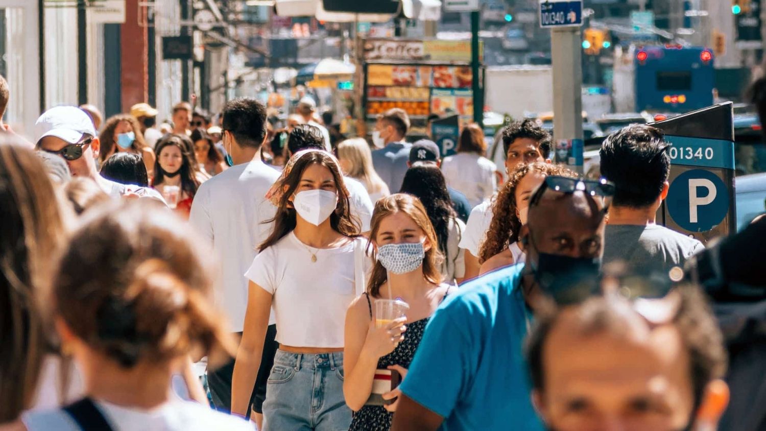 people of many races walk along a crowded street. some of them are wearing masks.