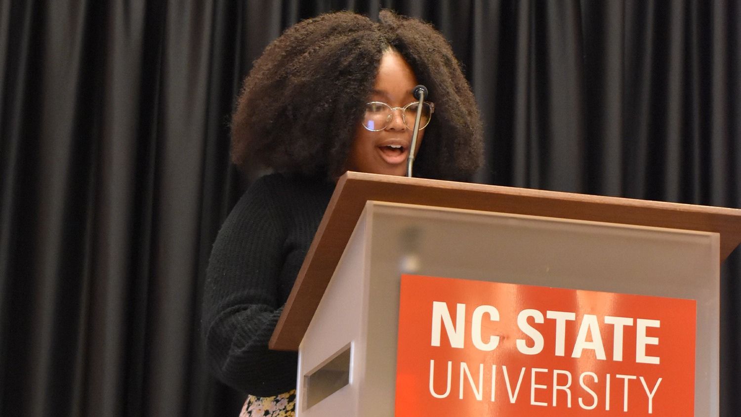 Naudia McKoy standing behind a podium with the NC State logo on it and giving a speech