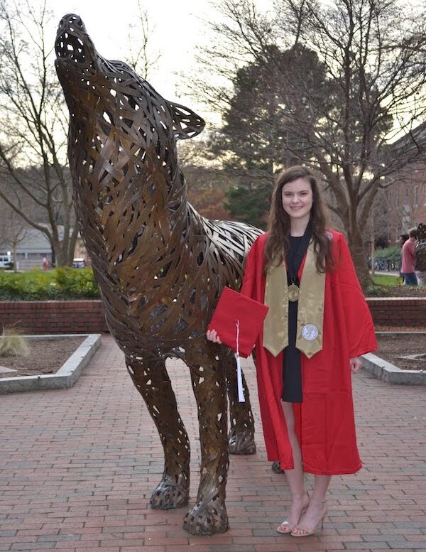 Jessica Parzygnat wearing an NC State graduation gown posing with a wolf statue