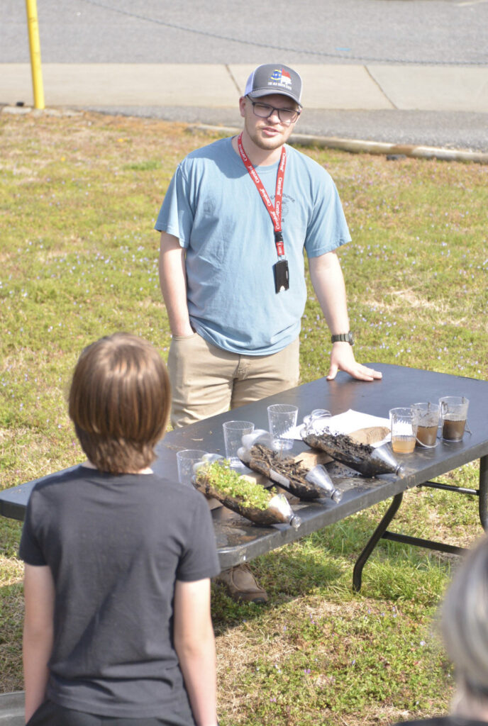 a man stands at a table explaining different plants and soils exhibited in cups and bottles.