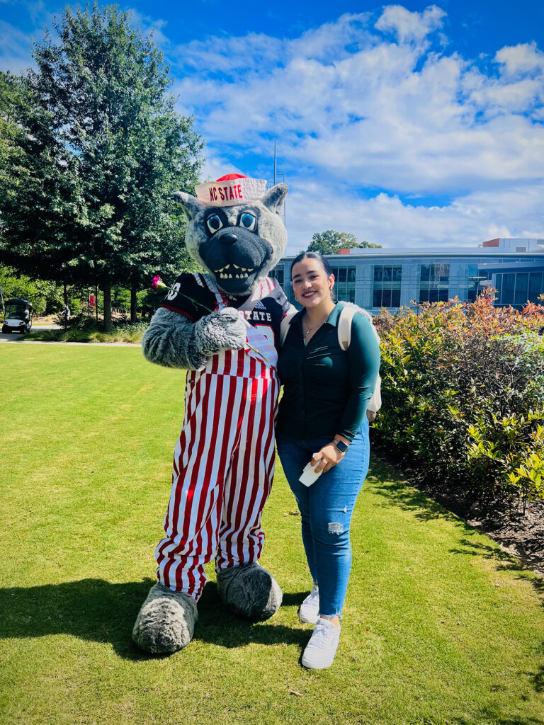 Young woman standing on a green lawn with a wolf mascot in striped overalls