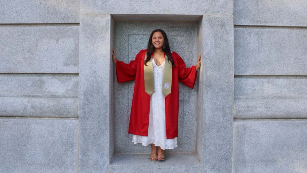 a woman wearing a red graduation gown stands by a gray marble wall