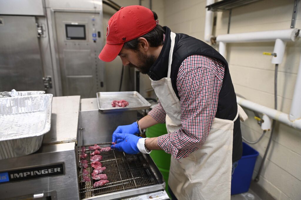 Man in red hat and white apron and blue gloves grills meat