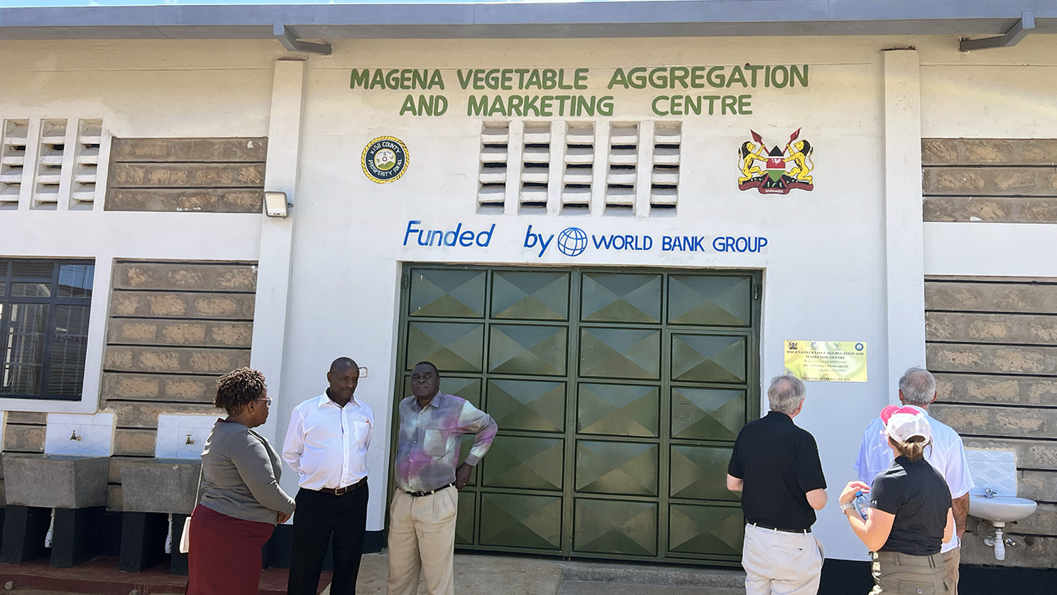 a group of people stand outside of a building with a sign that reads Megena Vegetable Aggregation and Marketing Centre