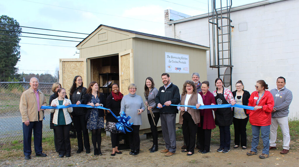 a group of people stand next to a small shed holding large blue ribbon and cutting it.