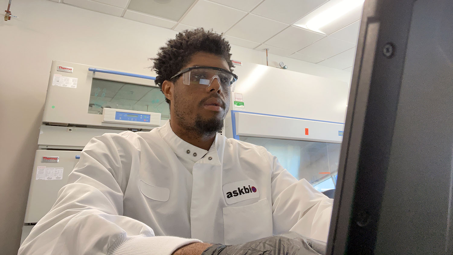 a man wearing a lab coat and safety glasses looks at a computer in a science lab