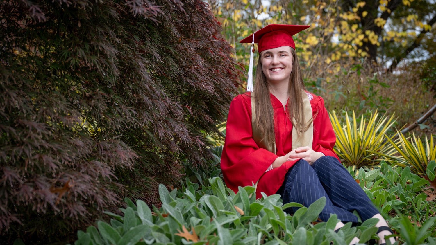 a woman wearing a red cap and gown sits among plants