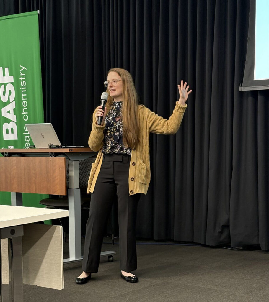 a women holds a microphone while making a presentation with a BASF sign in the background