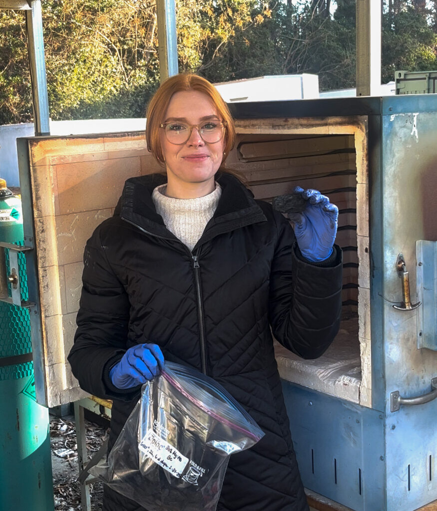 a woman stands in front of a kiln holding a black chunk of biochar