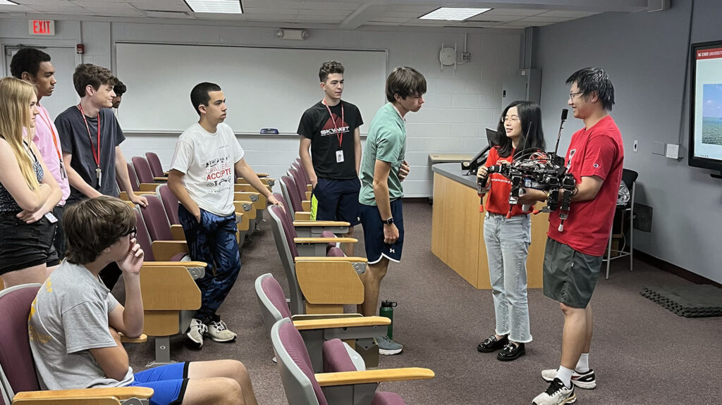 students in a classroom look at man and woman holding a spider-like robot