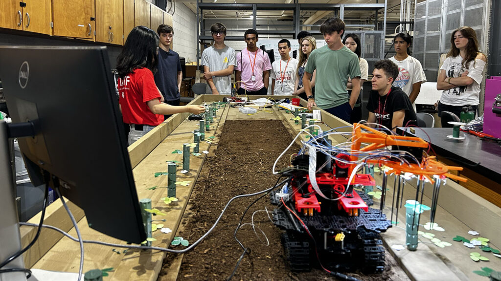 a group of students stand around a test dirt table looking at prototype robots