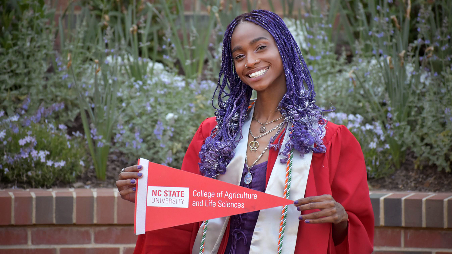 a woman wears a read graduation robe and holds a triangular sign that says 'NC State University College of Agriculture and Life Sciences'