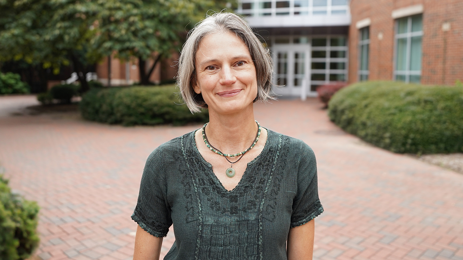 a woman with gray hair wearing a shortsleeved moss green shirt stands in front of a brick building