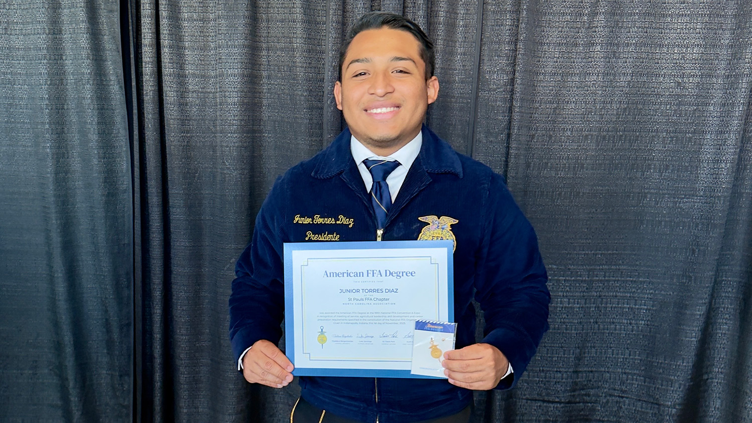 Young man in a blue blazer in front of gray curtain holding a certificate