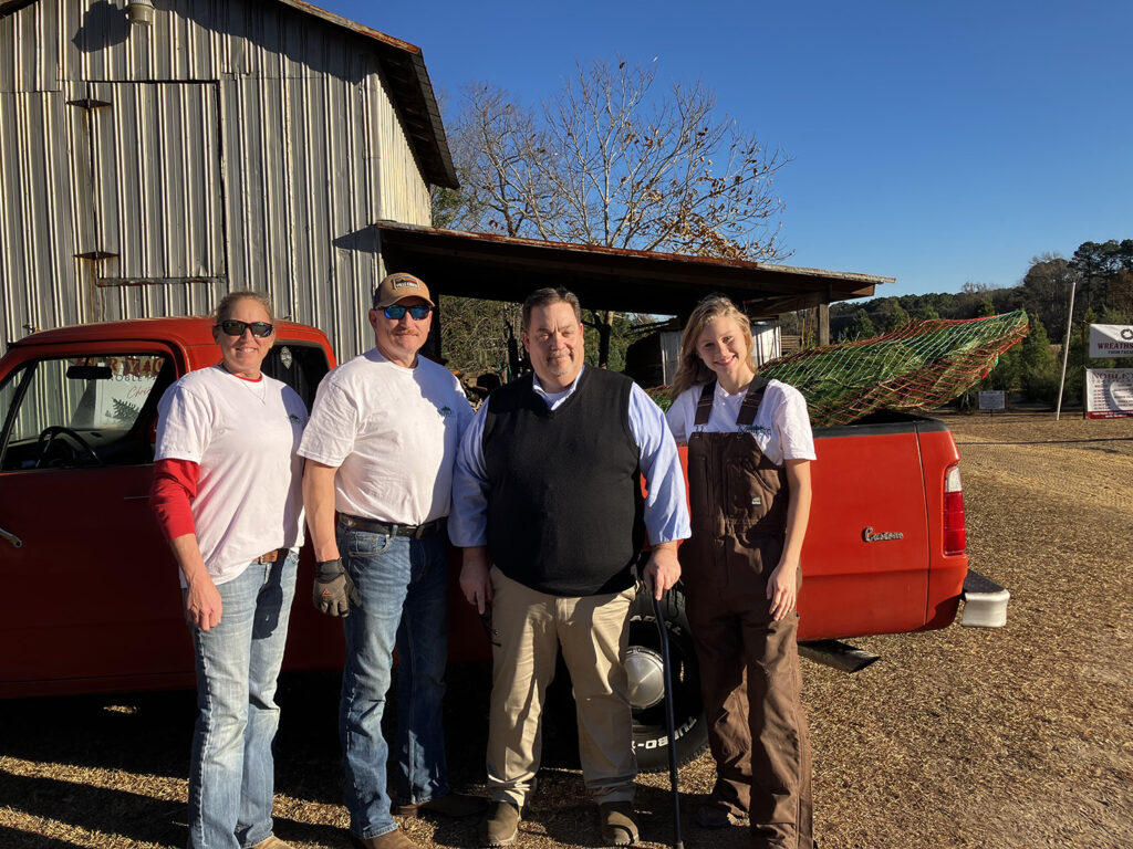 a group of people stand in front of a red pick up truck with a netted christmas tree in the back