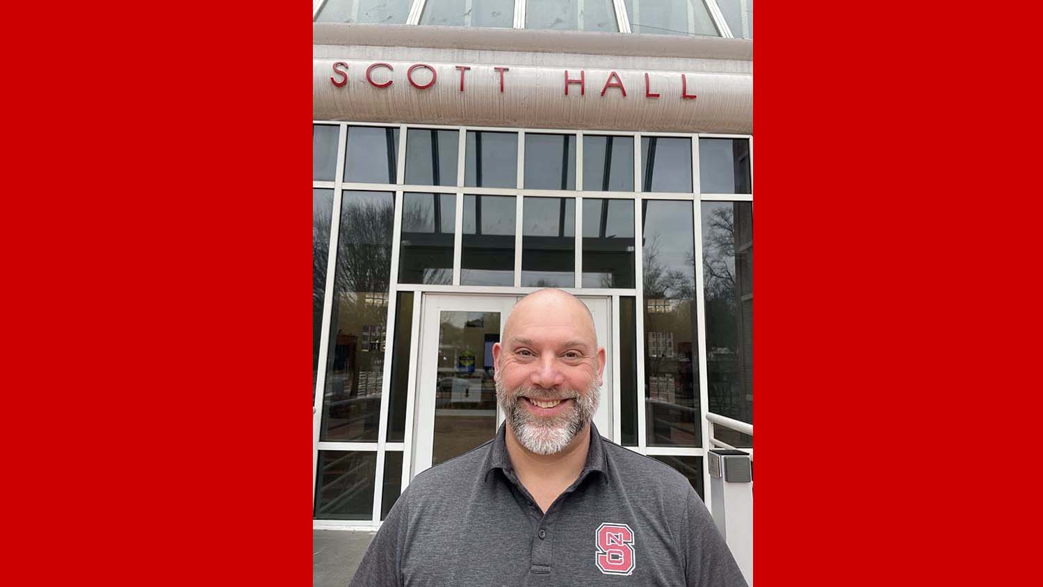 a man stands in front of a glass window building with the words Scott Hall above the door