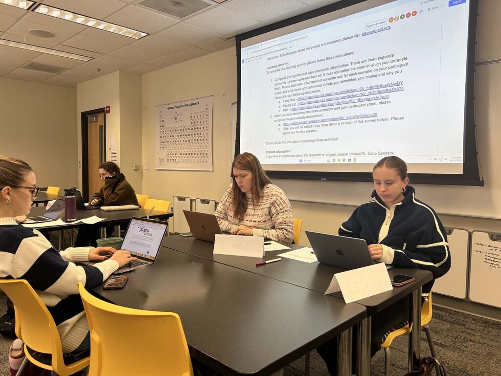 students sit at black tables while looking at laptops in a classroom with a whiteboard screen projector displaying instructions in the background