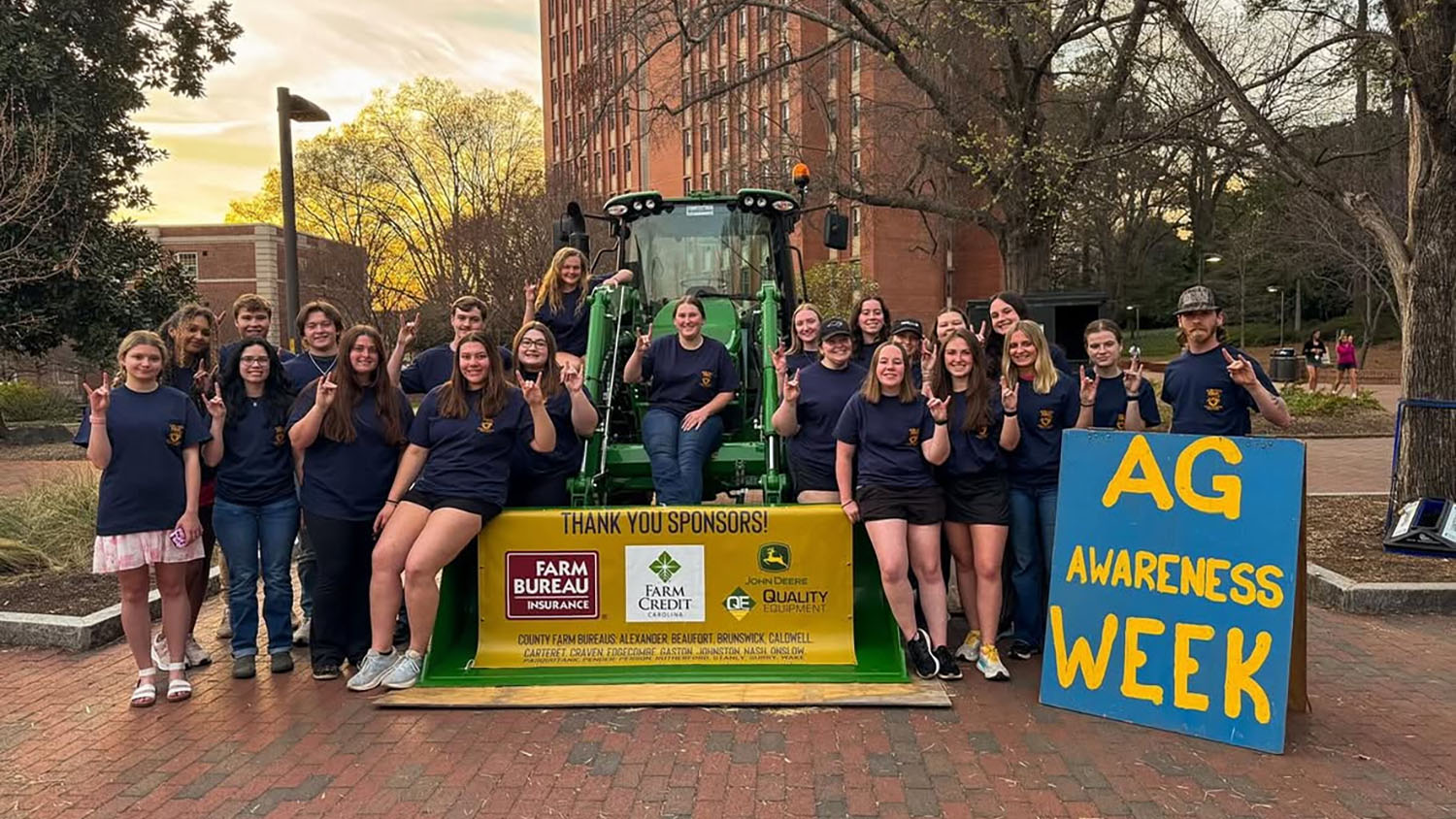 a group of people stand on a brick pathway with a tractor and a sign that says Ag Awareness Week