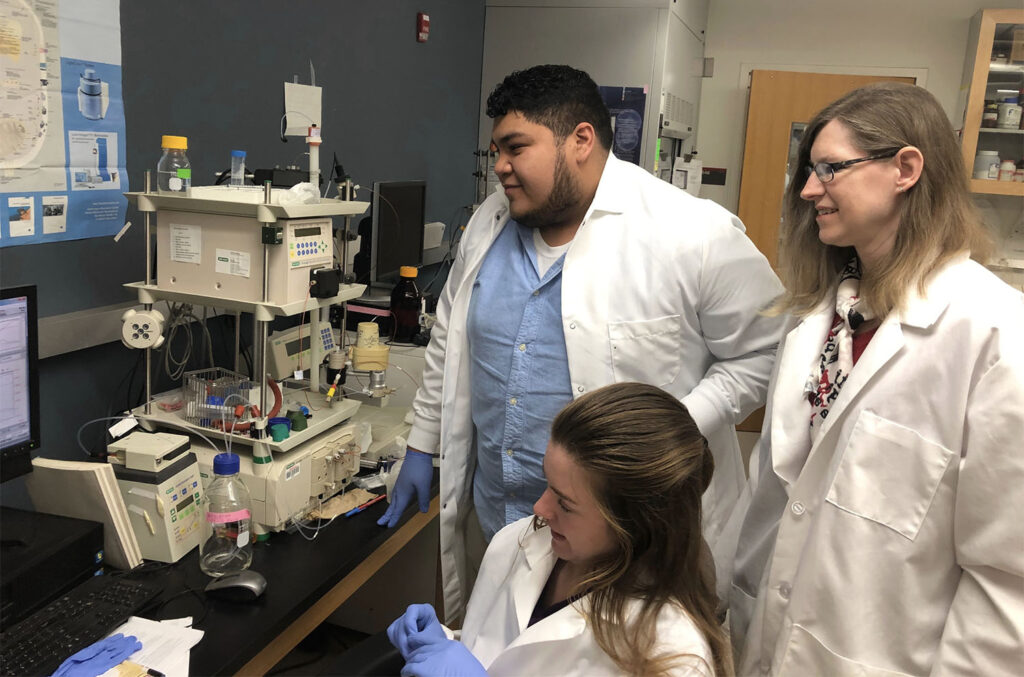 three people wearing white lab coats in a science lab working with science equipment and computers