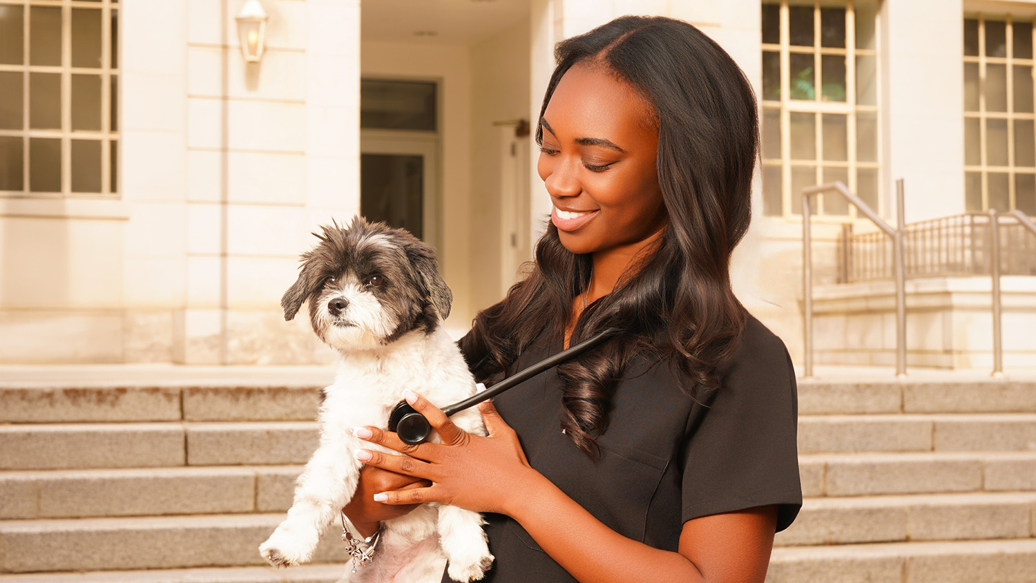 A young woman smiling, holding a stethoscope to a small black and white dog's chest, in front of a campus building's staircase.