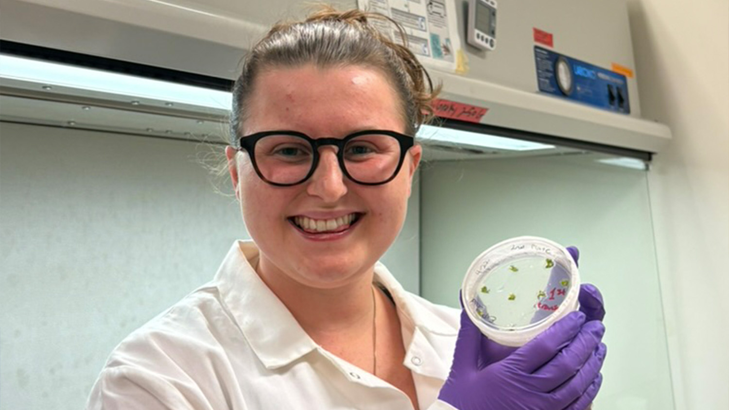 A woman wearing glasses and a lab coat smiling while holding a petri dish.