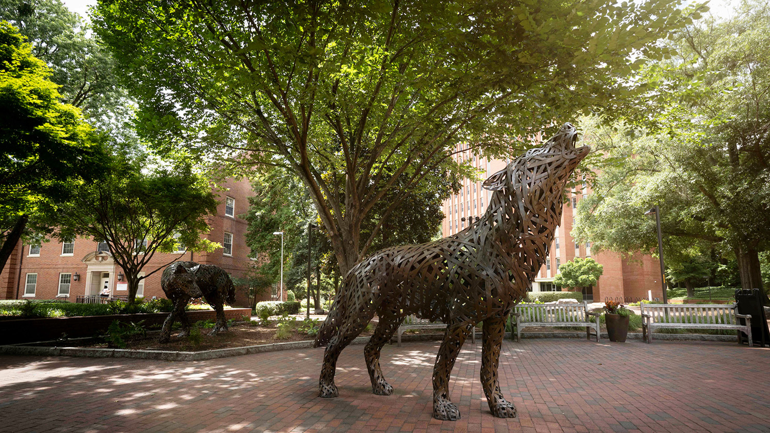 two wolf statues made of copper weaved metal standing on a brick patio surrounded by trees