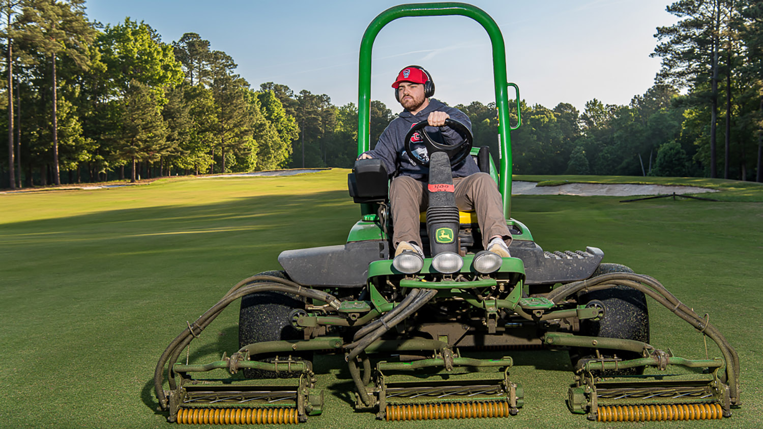 a man wearing a red baseball hat, grey sweatshirt and khaki pants drives a large industrial lawn mower on a golf course