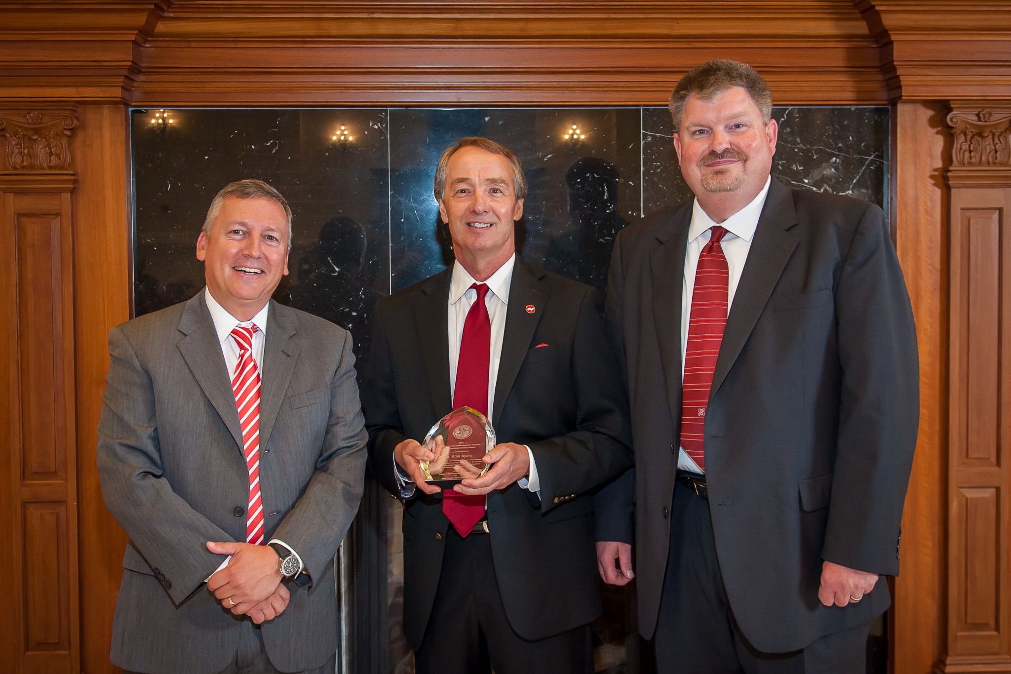 Three men standing in suits while the one in the middle is holding an award.