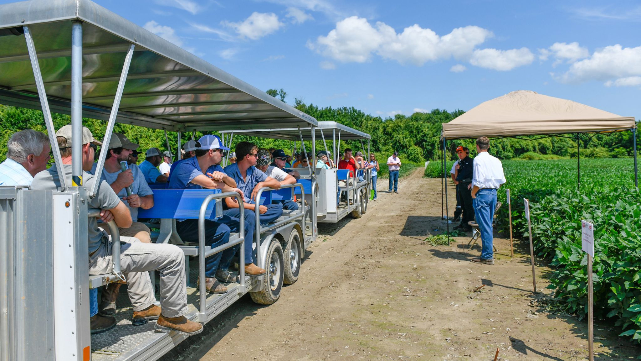 People riding a trailer-trolley stop amid a soybean field to listen to a man speak.